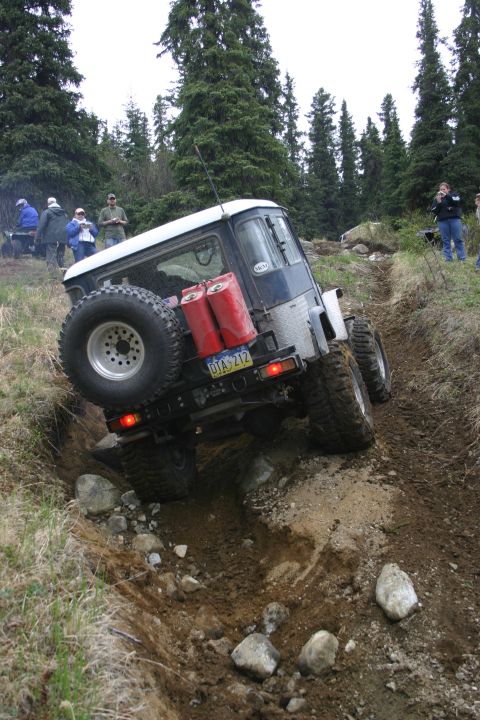 2003 Jeep TJ - An off-road vehicle navigates a challenging muddy and rocky terrain, equipped with a spare tire and auxiliary gas cans.