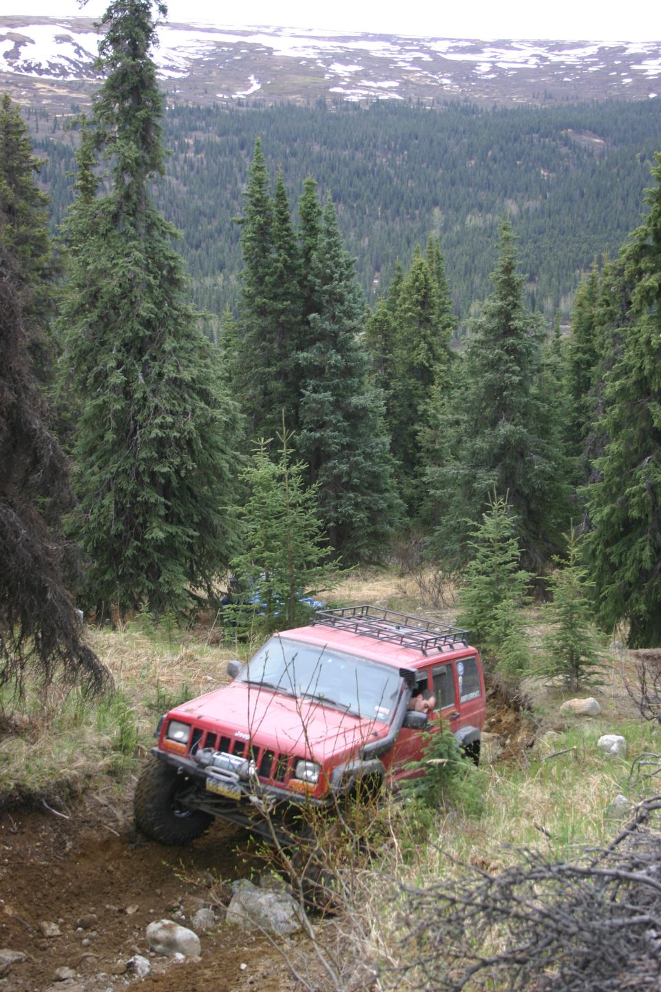 Red Jeep Cherokee XJ driving on a dirt trail through a forest with snow-capped mountains in the background