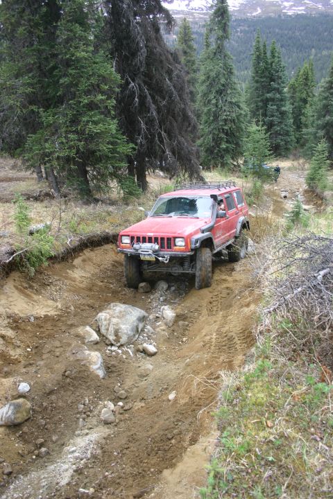 2003 Jeep TJ - A red Jeep Cherokee XJ navigates a rough, muddy trail surrounded by dense forest and distant mountains.