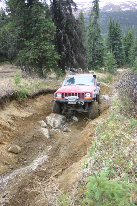 2003 Jeep TJ - A red Jeep Cherokee XJ navigates a challenging off-road trail with steep, muddy banks and scattered rocks.