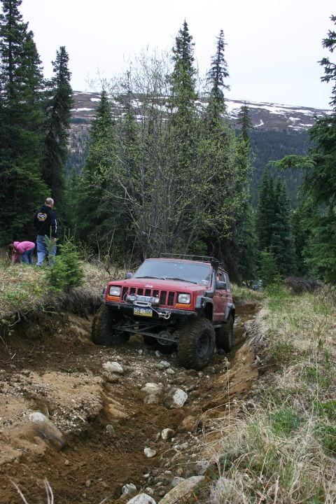 2003 Jeep TJ - Modified Jeep Cherokee XJ tackling a challenging off-road trail surrounded by trees and mountainous terrain.