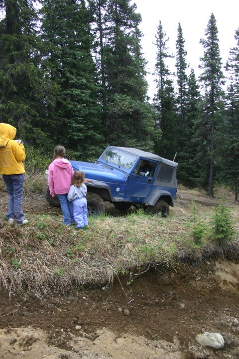 2003 Jeep TJ - A blue Jeep TJ is parked on a dirt path surrounded by a forest, with three people observing.