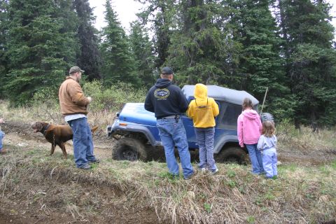 2003 Jeep TJ - A blue Jeep Wrangler is stuck in the mud with a group of people and a dog observing.