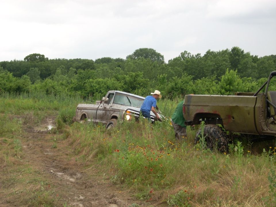 Two trucks stuck in a muddy field with people trying to free one
