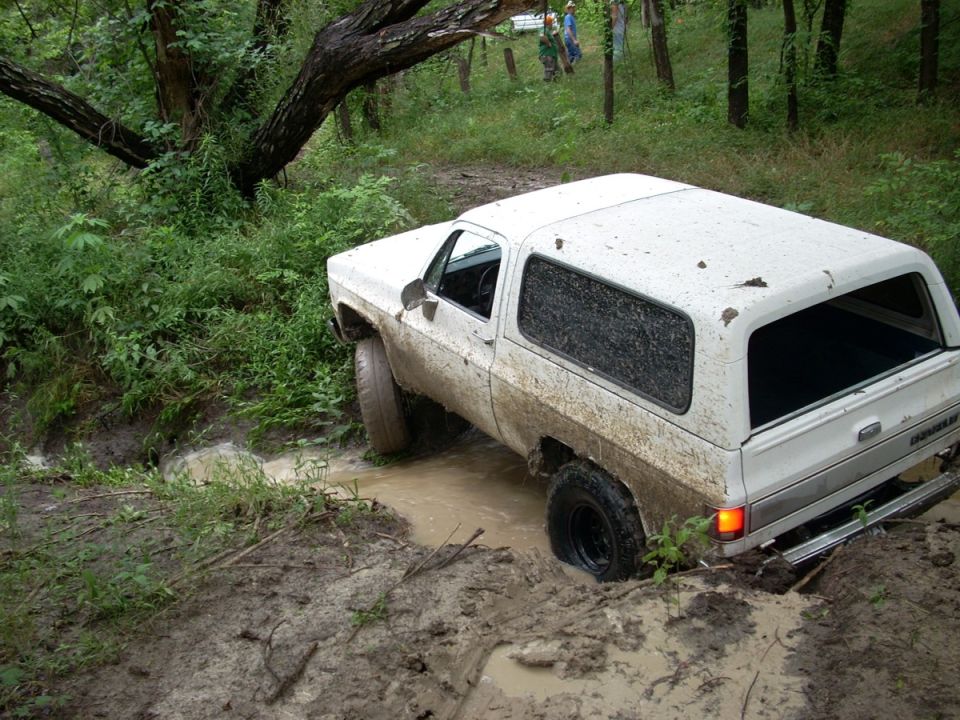 White full-size SUV stuck nose-down in a muddy creek crossing in the woods, covered in mud
