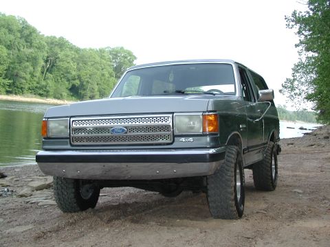 1989 Ford Bronco - This Ford Bronco is parked on a dirt bank next to a body of water, with trees in the background.