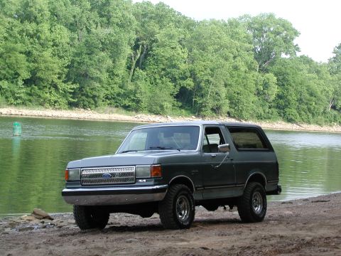 1989 Ford Bronco - This Ford Bronco is parked on a sandy shore next to a body of water with a wooded area behind it.