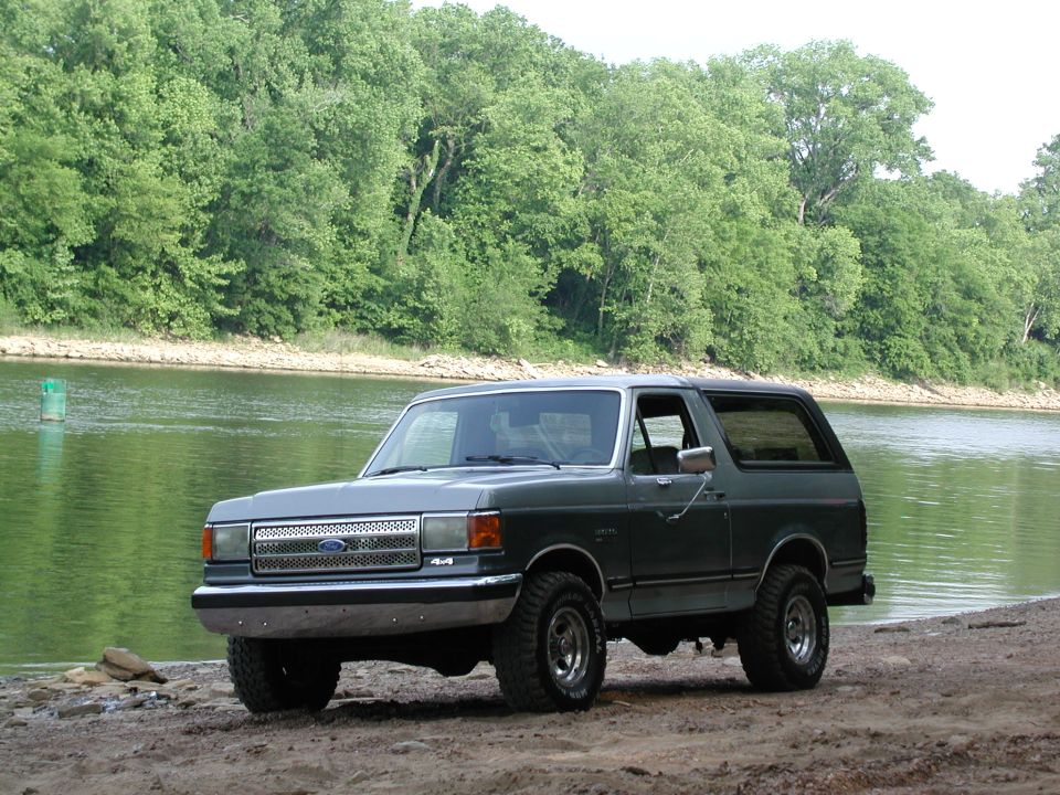 Gray Ford Bronco parked by a river with trees in the background