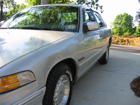 1985 Ford LTD Crown Victoria - Low front-quarter close-up of a silver Ford Crown Victoria four-door sedan parked on a concrete driveway, showing the clear headlight with…