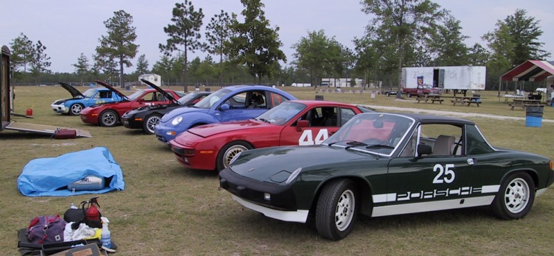 Dark green Porsche 914 with number 25 leading a row of race-numbered cars parked on grass at a paddock.