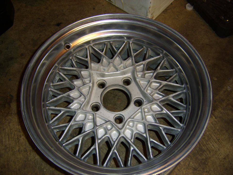 Bare silver lattice-mesh alloy wheel with machined lip and five-lug hub on a shop floor