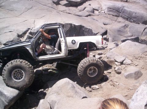 1989 Ford Bronco - Passenger-side view of a white doorless mini-truck rock crawler flexing over rough granite, with a tube exo-cage, red coilover shocks, high…