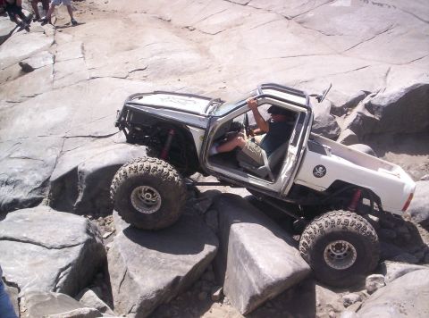 1989 Ford Bronco - A white doorless mini-truck rock crawler with an exo-cage, coilover shocks and a Jolly Roger decal on the bedside climbs a granite slab, dr…
