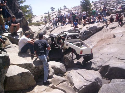 1989 Ford Bronco - White doorless mini-truck rock crawler dropping through a granite obstacle, showing its exo-cage, red coilover shocks, skull-and-crossbones…
