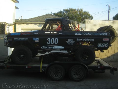 1990 Ford Bronco - black off-road race truck numbered 300 lettered 'West Coast Broncos' with sponsor decals sits chained on a flatbed trailer.