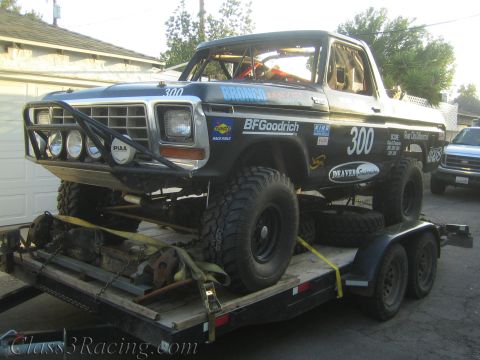 1990 Ford Bronco - Front view of a black off-road race truck number 300 with a chrome grille, light bar and brush guard strapped on a trailer.