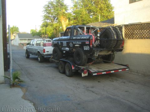 1990 Ford Bronco - black off-road race truck marked 'West Coast Broncos' sits strapped on a tandem-axle flatbed trailer towed by a silver Ford pickup in a nar…