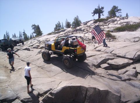 1989 Ford Bronco - Off-road vehicles navigate a rocky terrain under a clear blue sky, with one vehicle carrying an American flag.