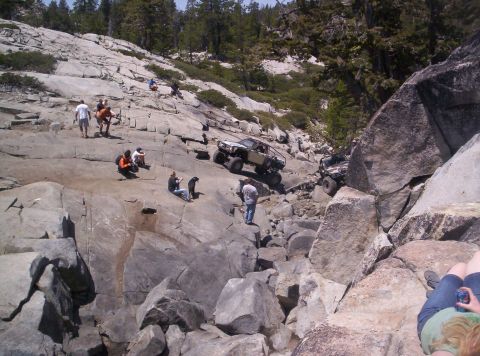 1989 Ford Bronco - People watch as modified off-road vehicles tackle a challenging rocky incline in a natural landscape.