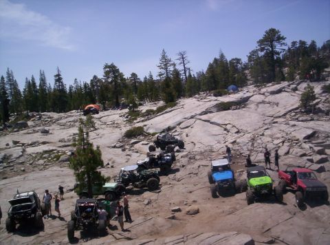 1989 Ford Bronco - Several off-road vehicles are gathered on a rocky terrain with trees and tents in the background.