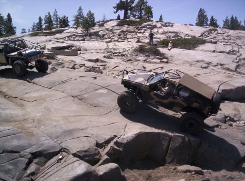 1989 Ford Bronco - Two modified off-road vehicles navigate a challenging rocky incline, one assisting the other with a tow rope.