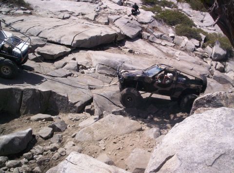 1989 Ford Bronco - An off-road vehicle navigates a rocky terrain, assisted by a tow rope from another vehicle.