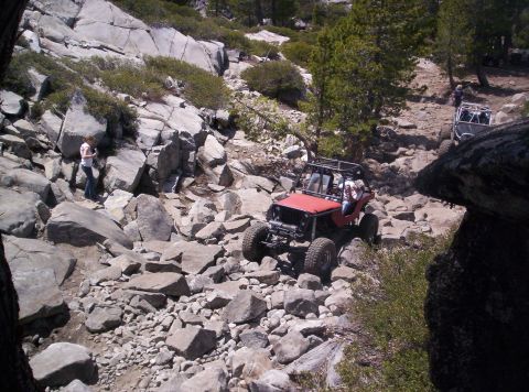 1989 Ford Bronco - Overhead view down a granite boulder trail as a red flat-fender-style Jeep buggy with a full roll cage, tube bumper and oversized tires wor…