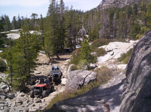 1989 Ford Bronco - Several off-road vehicles traverse a challenging, rocky path through a wooded mountain landscape.