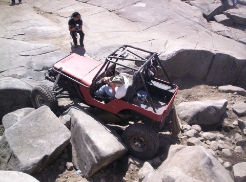 1989 Ford Bronco - A red off-road jeep is shown maneuvering through a challenging rocky landscape with a person observing in the background.