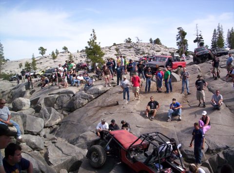 1989 Ford Bronco - A large group of people and several off-road vehicles are gathered on a rocky, mountainous landscape under a clear blue sky.
