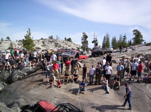 1989 Ford Bronco - A group gathers around off-road vehicles parked on a rocky, mountainous terrain under a clear blue sky.