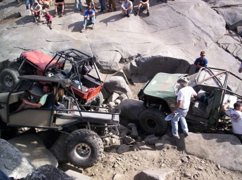 1989 Ford Bronco - Multiple off-road vehicles are engaged in a challenging rock crawling event with an audience observing from a higher elevation.