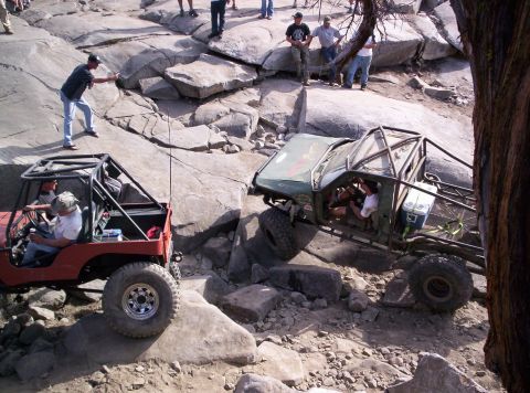 1989 Ford Bronco - Two heavily modified off-road vehicles traverse a challenging rocky course with spectators watching.