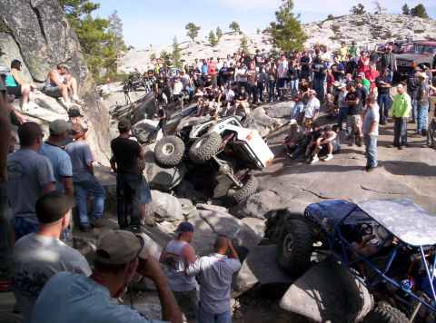 1989 Ford Bronco - A crowd gathers to watch an off-road vehicle navigate a challenging rocky terrain, with one vehicle overturned.