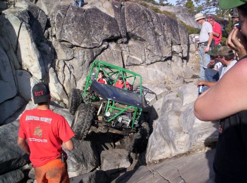 1989 Ford Bronco - An off-road buggy with a green roll cage is being driven over large rocks, with spectators observing the side.