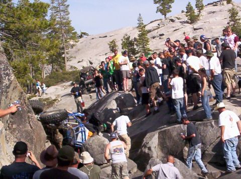 1989 Ford Bronco - A crowd gathers to watch an off-road vehicle that has flipped onto its side on a rocky terrain.