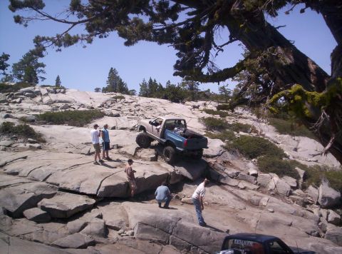 1989 Ford Bronco - A group of people observe an off-road pickup truck navigating a challenging rocky incline on a sunny day.