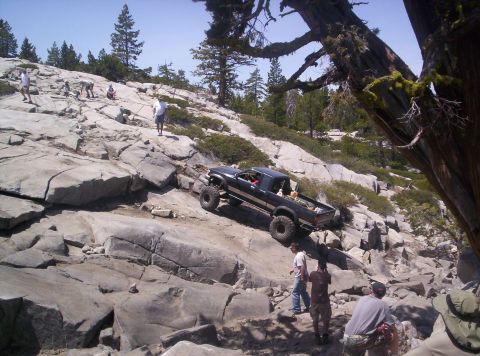 1989 Ford Bronco - A modified black pickup truck tackles a challenging off-road course on a sunny day, with several onlookers observing the ascent.