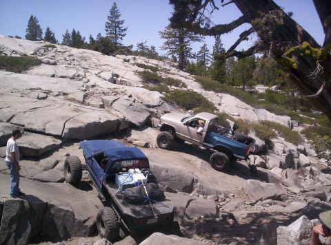 1989 Ford Bronco - Two modified trucks tackle a challenging rocky trail in a mountainous, wooded area.