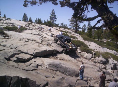 1989 Ford Bronco - A black pickup truck navigates a challenging rocky terrain, with onlookers observing the off-road adventure.