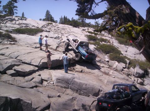1989 Ford Bronco - Two modified trucks navigate a challenging rocky terrain on a sunny day.