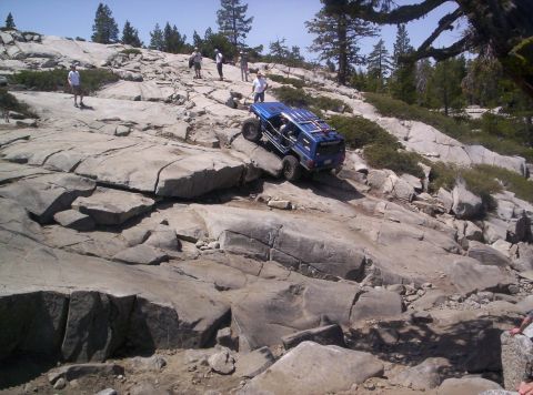1989 Ford Bronco - A blue off-road SUV tackles a challenging rocky terrain, with several onlookers observing the ascent.
