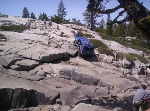 1989 Ford Bronco - A blue off-road SUV navigates a challenging rocky path, with onlookers observing the ascent.