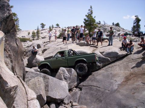1989 Ford Bronco - A group of spectators watches as a green pickup truck maneuvers over large rocks on an off-road trail.
