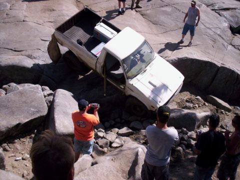 1989 Ford Bronco - A white pickup truck is precariously positioned on large rocks, with several onlookers observing the scene.