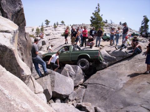 1989 Ford Bronco - A group of people watch as a green pickup truck attempts to maneuver over large rocks on an off-road trail.