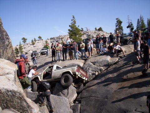 1989 Ford Bronco - A group of people watch as a Toyota pickup truck attempts to climb over large rocks on a sunny day.