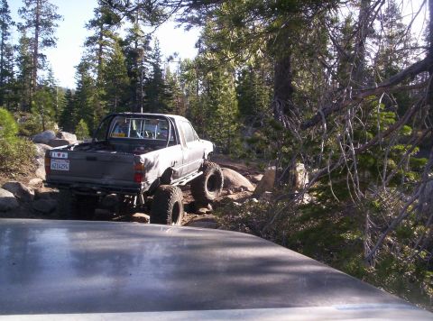 1989 Ford Bronco - A lifted pickup truck navigates a challenging rocky trail through a forest, with another vehicle visible in the foreground.