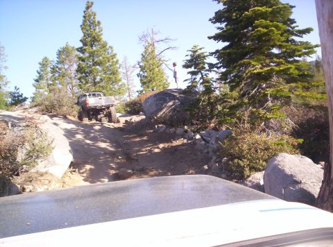 1989 Ford Bronco - A silver pickup truck navigates a rocky off-road trail, with another vehicle's hood in the foreground.