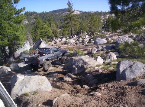 1989 Ford Bronco - A modified pickup truck tackles a challenging rocky terrain in a wooded, mountainous area.